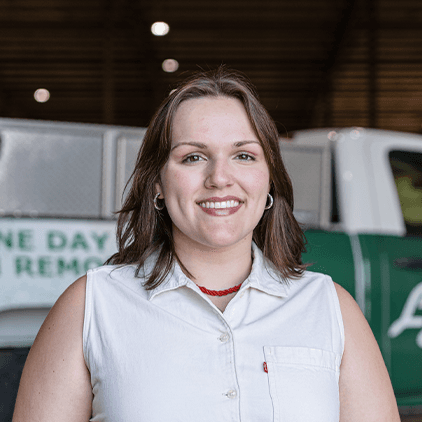 Woman with shoulder-length brown hair smiling at camera, wearing white sleeveless button-up shirt in front of industrial building interior