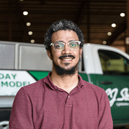 Man wearing maroon polo shirt and glasses smiling at camera in front of green and white work trucks in warehouse setting