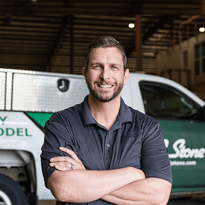 Smiling man in dark polo shirt with arms crossed, standing in front of white and green service vehicles in a garage