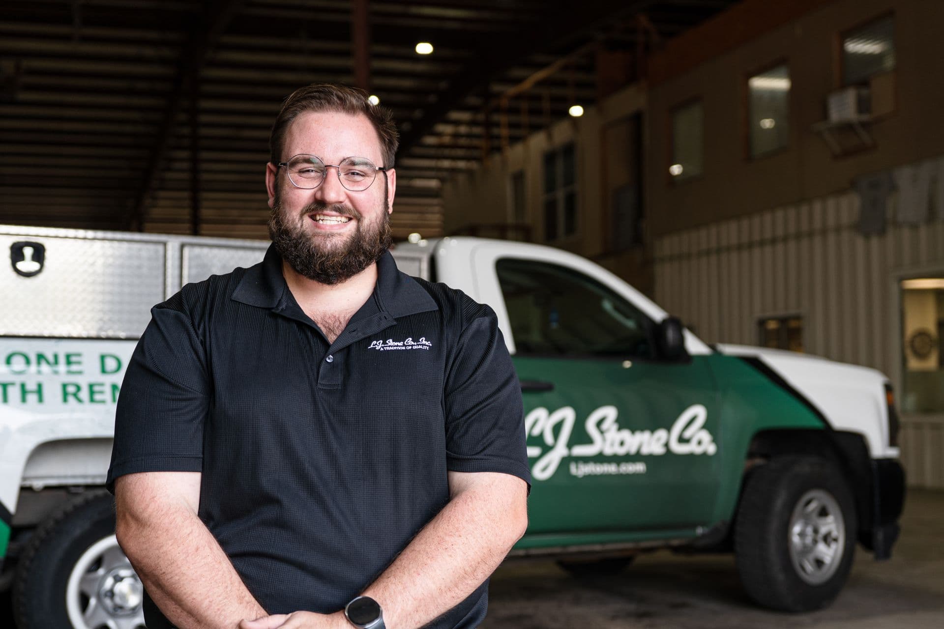 Man wearing glasses and navy polo shirt smiling at camera in front of green and white company service vehicles in warehouse setting