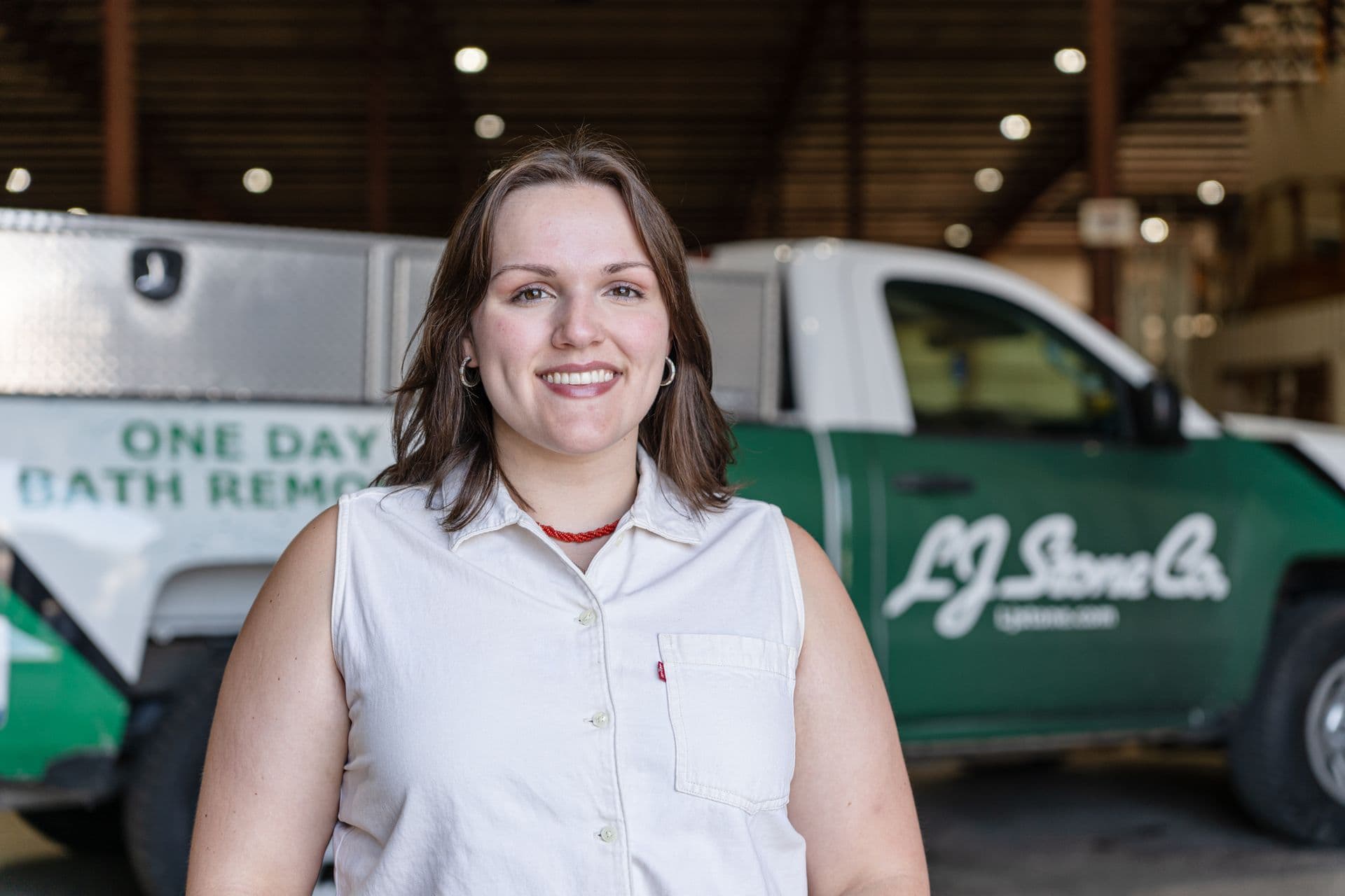 Woman in white sleeveless work shirt smiling at camera in front of green and white service vehicles in a garage