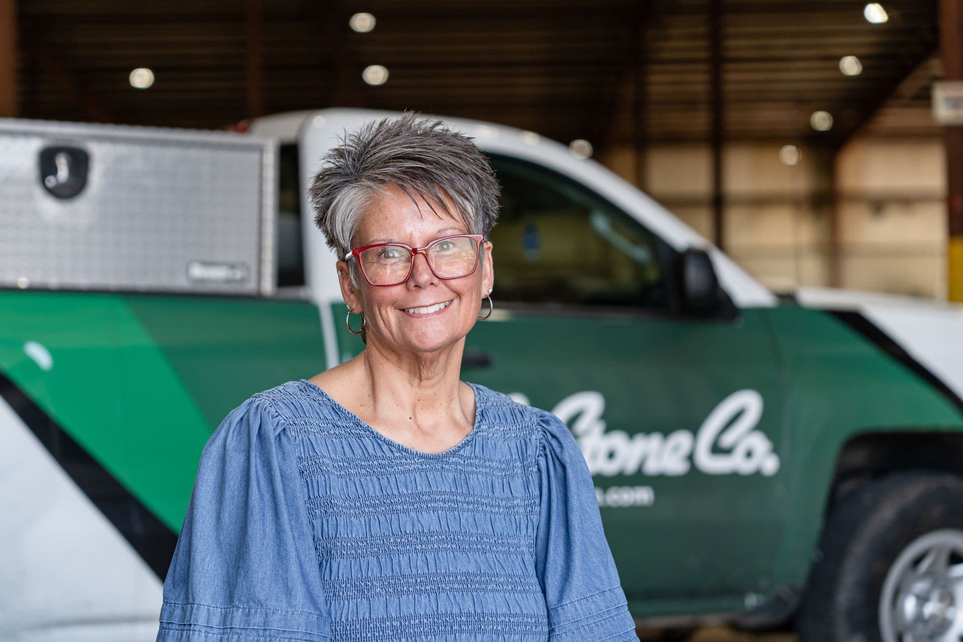 Woman with short gray hair and red glasses smiling in front of a green and white service vehicle in a garage