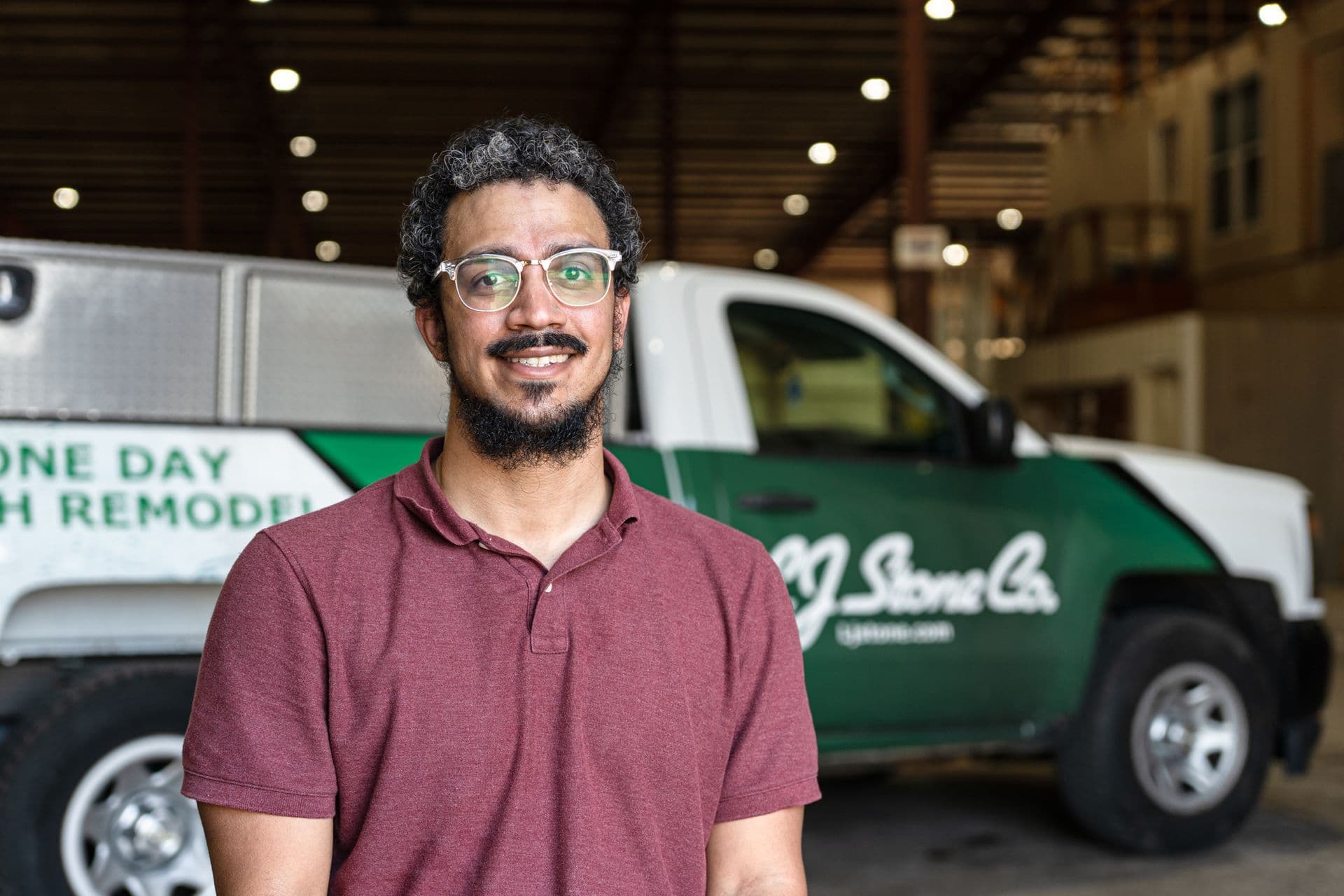 Man wearing glasses and maroon polo shirt smiling at camera in front of green and white service vehicles in a garage