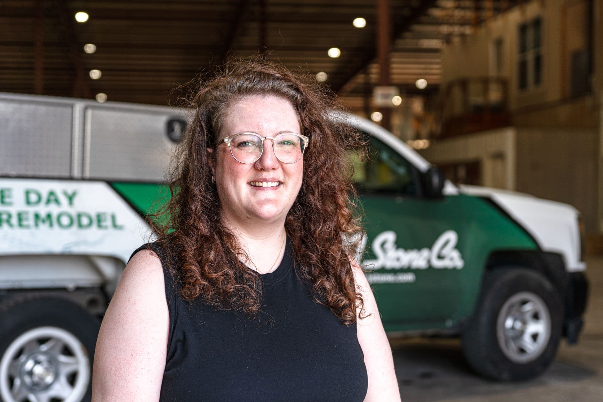 Smiling woman with curly brown hair and glasses standing in front of a green and white service truck in a garage