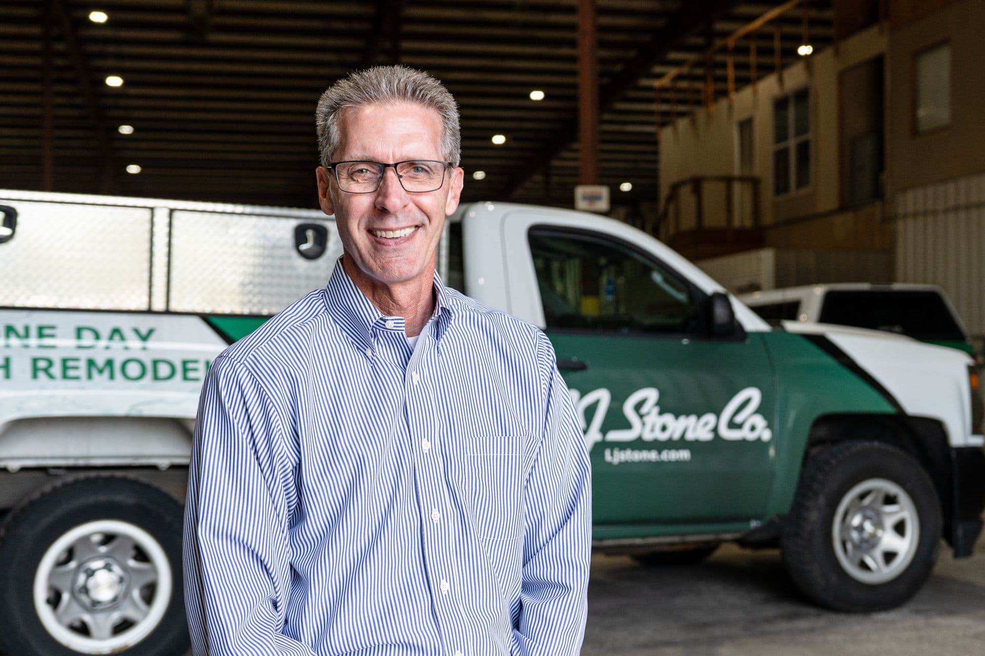 Man in blue striped shirt smiling in front of company service vehicles in warehouse