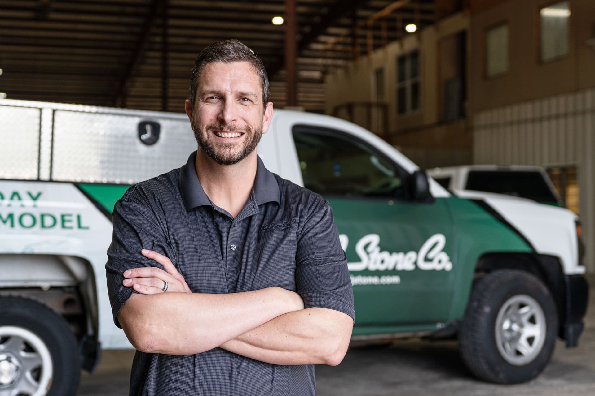 Professional man with arms crossed smiling at camera in front of green and white company service trucks in warehouse facility