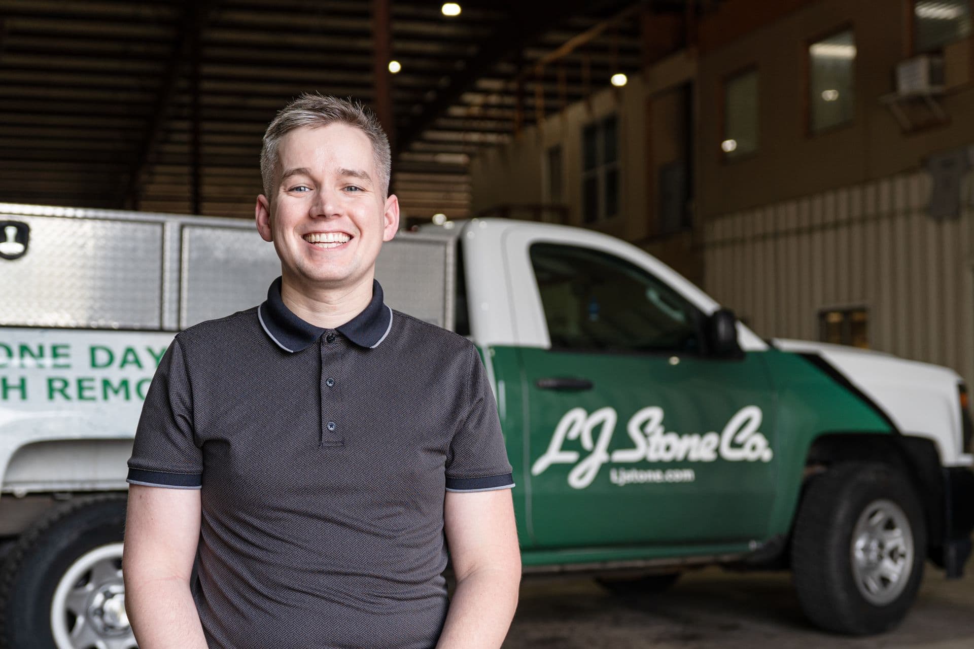 Smiling man in dark polo shirt standing in front of LJ Stone Co service vehicles in a warehouse garage