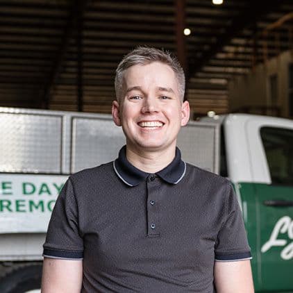 Smiling man wearing dark polo shirt standing in front of white and green work vehicles in a warehouse setting