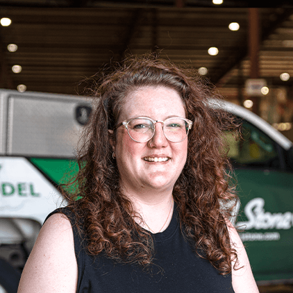 Person with long curly brown hair and glasses smiling at camera indoors near vehicles