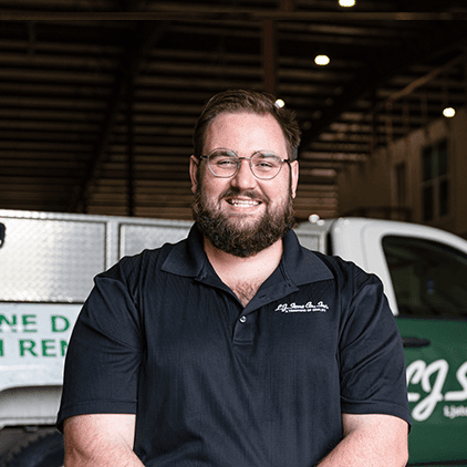 Man in dark polo shirt standing in front of vehicles in a garage or warehouse setting, smiling at the camera